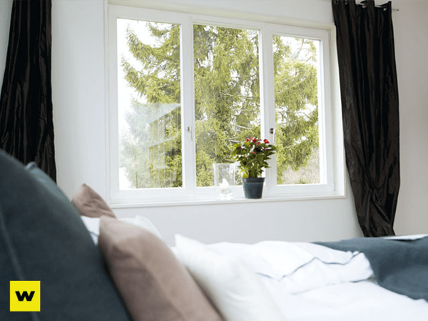 Helles Schlafzimmer mit großem Fenster, Bett und Zimmerpflanze, gemütliche Wohnatmosphäre.