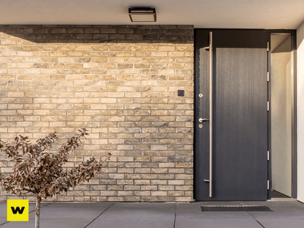 Moderne Haustür aus dunklem Metall neben heller Klinkerfassade mit Pflanze im Eingangsbereich.